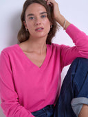 A woman wearing a bright pink long-sleeve V-neck knit top and dark blue jeans, posing against a plain light background.