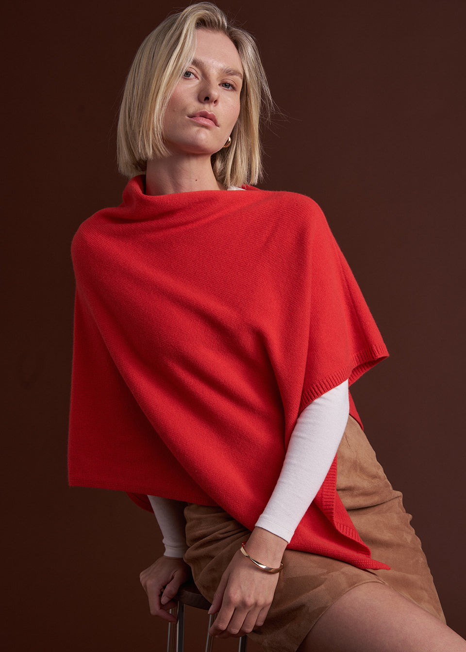 A woman wearing a bright red knit poncho draped over her shoulders, layered over a white long-sleeve top and tan skirt, posed against a dark brown studio background.