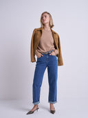 A woman modeling a tan suede jacket, beige knit top, blue jeans, and brown pointed-toe heels against a light gray studio background.