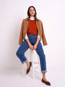 A woman models a brown suede jacket over a rust-orange turtleneck and blue jeans, sitting on a white stool against a plain white studio background.