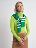 A woman wearing a bright lime green long-sleeve turtleneck sweater and a green and white striped knit scarf, paired with a brown skirt, against a light gray studio background.