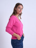 A woman wearing a bright pink long-sleeve V-neck sweater and dark blue jeans, standing against a light gray studio background.