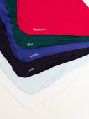 Five folded fabric scarves or bandanas in different colors are stacked diagonally on a white surface. The top scarf is bright red with the name 'Raspberry' printed in white, followed by dark green 'Pine', blue 'Lobelia', dark navy 'Indigo', and very pale blue 'White'.