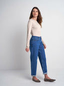 A woman modeling a pair of high-waisted, straight-leg blue jeans with rolled cuffs, paired with a beige long-sleeve top and brown flats, set against a plain light gray studio background.