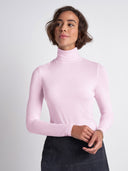 A woman wearing a light pink long-sleeve mock neck top and a dark skirt, standing against a light gray studio background.