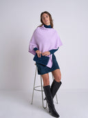 A woman models a lavender and navy blue color-block poncho sweater, styled with a matching navy clutch, a navy mini skirt, and black knee-high boots, while sitting on a metal stool against a light gray studio background.