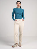 A woman modeling a teal and navy blue striped long-sleeve knit top with cream-colored trousers and beige sneakers, standing against a light gray studio background.