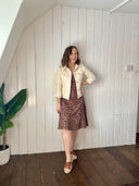 Woman standing in a room with white paneled walls and wooden floor, wearing a cream jacket and patterned dress.