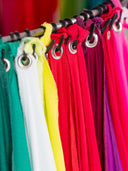 A close-up of a clothing rack displaying multiple colorful fabric panels or curtains in green, white, yellow, red, magenta, and purple, each attached with a black grommet to a metal rod.