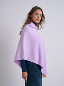 A woman wearing a light purple/lavender cowl-neck poncho over a dark navy long-sleeve top and navy skirt, photographed against a neutral light gray studio background.