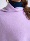 Close-up of a person wearing a soft lavender knit cowl-neck sweater or poncho over a dark navy turtleneck, with brown hair visible against a plain light background.