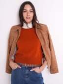 A woman wearing a tan suede jacket over an orange crewneck sweater, a white turtleneck, and blue jeans, standing against a plain white background.