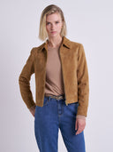 A woman modeling a cropped tan suede jacket over a beige top and blue jeans, photographed against a plain light gray studio background.