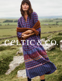 A woman standing outdoors in a grassy landscape wearing a loose, long-sleeve plaid dress in blue, red, and purple tones, with a brown wrap draped over her arms. The brand name 'CELTIC & CO.