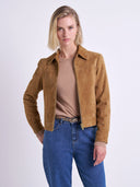 A woman modeling a cropped tan suede jacket over a beige top and blue jeans, photographed against a plain light gray studio background.