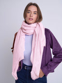 A woman modeling a light pink fringed scarf wrapped around her neck, paired with a purple jacket, light blue top, and dark blue jeans against a plain light gray studio background.