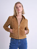 A woman wearing a cropped tan suede jacket with a front zipper, paired with a beige top and blue jeans, photographed against a light gray studio background.