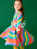 A young girl with long brown hair is wearing a colorful, sleeveless A-line dress with wavy rainbow stripes in pink, orange, yellow, green, blue, and purple. She is standing against a solid dark green studio background, smiling and twirling.