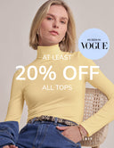 A blonde woman wearing a fitted pale yellow mock-neck long-sleeve top and blue jeans sits on a woven chair against a light beige studio background. Large text reads 'AT LEAST 20% OFF ALL TOPS,' with a light blue circle badge that says 'AS SEEN IN VOGUE.