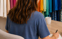 A woman with long brown hair is seen from behind, wearing a slate blue t-shirt and sitting at a wooden table, writing in a notebook. Colorful clothing racks are visible in the background.