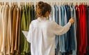 A woman viewed from behind stands in front of a rack of long-sleeve shirts, touching a blue one while holding a tablet. The shirts are displayed in a variety of colors including beige, brown, green, blue, and red.