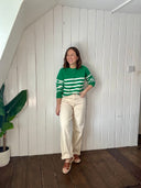 Person wearing a green and white striped sweater and beige pants standing against a white paneled wall.